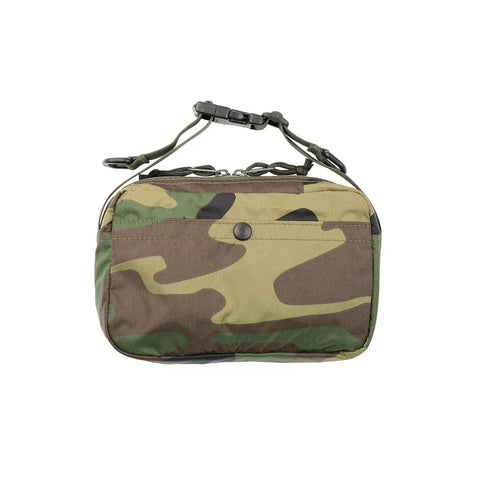 The MIS AW Shoulder Bag S in Woodland Camo is a small rectangular packcloth nylon bag with zippered top, front snap pocket, short buckle handle, and adjustable shoulder strap.