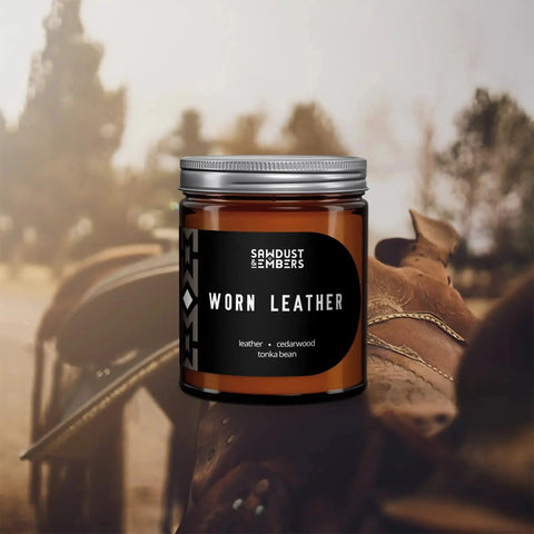 A brown glass jar of Sawdust & Embers Worn Leather - Signature Candle is displayed before a blurred background featuring a leather saddle and trees. Its label highlights leather, cedarwood, and tonka bean scents.