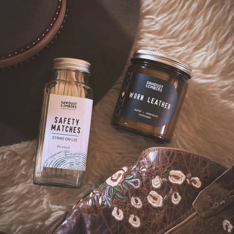 A jar of safety matches and a Sawdust & Embers Worn Leather - Signature Candle rest on a fuzzy surface beside a brown hat and floral-embroidered boots—perfect for adding American-made warmth to your space.