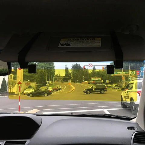 Seen from inside a car, a Glare Guard Yellow Polarized Car Visor Extender is attached to the sun visor, reducing glare. The clear street ahead features traffic lights and a police car parked on the right side of the road.