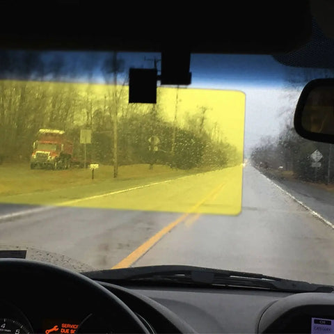 A view from inside a car shows the Glare Guard Yellow Polarized Car Visor Extender in use, making the left side of a rainy road look brighter and clearer than the dull right, with improved contrast and visibility. A red truck is on the left roadside.