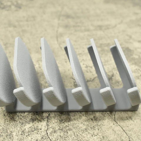 A close-up of the Chatelet Manufacturing Yeti Lid Mount Rack in gray, featuring five evenly spaced vertical slots, sitting on a textured beige surface. Ideal for efficient kitchen lid storage.