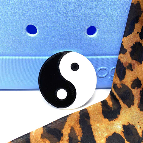 The BOGLETS Yin and Yang Charm by Chatelet Manufacturing, compatible with Bogg Bags, Simply Southern, and similar totes, sits on a textured blue surface beside bold leopard print fabric—ideal for personalizing your favorite bag.
