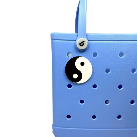 A close-up of a light blue tote bag featuring the Chatelet Manufacturing BOGLETS Yin and Yang Charm, compatible with Bogg Bags, Simply Southern, and similar totes, plus a small round button with the letter b on the strap.