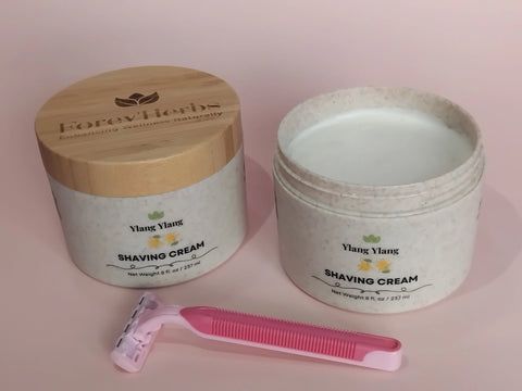 Two Forev'Herbs Ylang Ylang Shaving Cream containers, one with a wooden lid and one open to show the cream, are displayed beside a pink disposable razor on a light pink background.