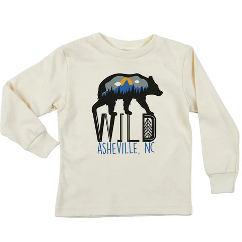 The Wild Bear Long Sleeve Graphic Tee by Spiritex | Sustainable Apparel is a cream youth shirt with a bear silhouette filled with mountains, trees, and sun, plus WILD ASHEVILLE, NC in bold black and blue for a fun organic kids design.