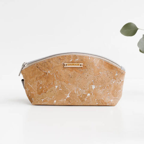 A tan, marbled cork zippered pouch—Carry Courage RULE BREAKER | SILVER—rests on a white surface, metal plate labeled COURAGE visible. Minimalist backdrop with a green leafy plant partially in view on the right.