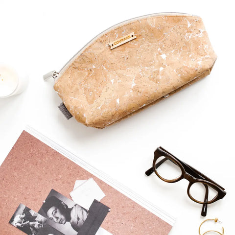 A silver, artisan-crafted RULE BREAKER zippered pouch by Carry Courage rests on a white surface beside black eyeglasses, a corkboard with black-and-white photos, a candle, and gold rings.