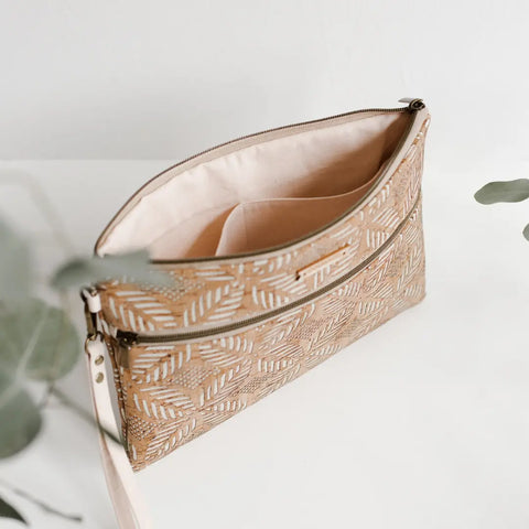 The Carry Courage INVENTOR tablet organizer in cream, crafted sustainably, features a leaf pattern, light pink interior, and inner pocket. It sits on a white surface accented with green leaves.