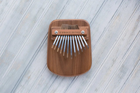 The Mountain Melodies Walnut 12 Key Thumb Piano is an American-made, handcrafted instrument with metal tines and numbered notes, set on a light whitewashed wooden base.