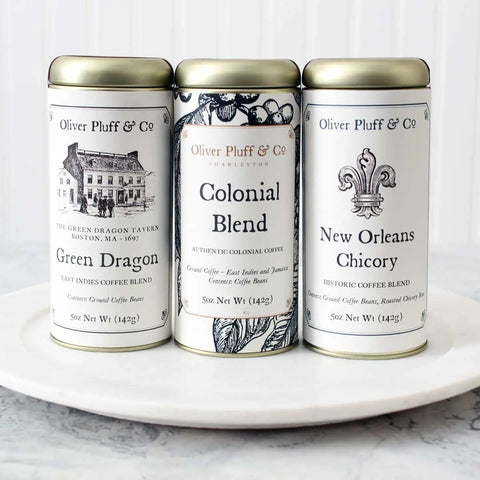 Three tins from the Early American Coffee Trio by Oliver Pluff & Company—Green Dragon, Colonial Blend, and New Orleans Chicory—feature vintage-style labels with black text celebrating America’s historic coffee culture on a white plate.