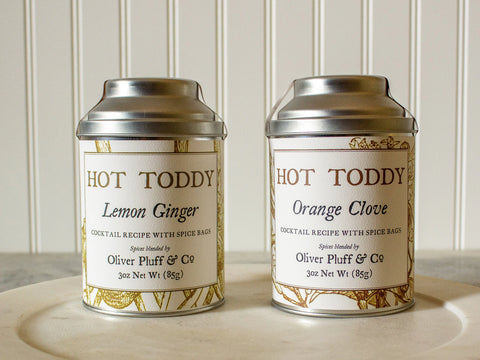 Two tins of Lemon Ginger and Orange Clove spices from Oliver Pluff & Company’s Your Hot Toddy Gift Set- A Soothing Tonic rest on a light surface with a white paneled background, perfect for making cozy bourbon drinks.