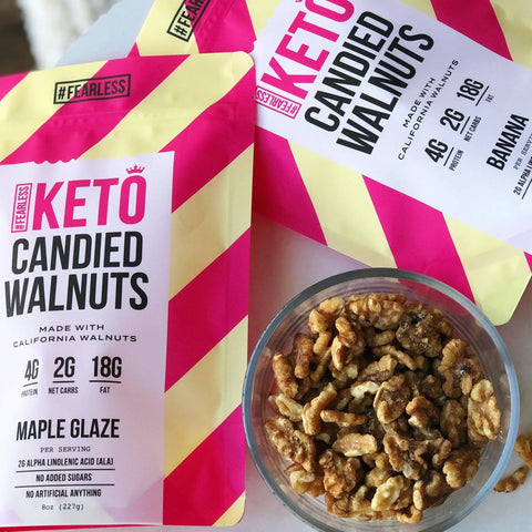 A bowl of walnuts sits beside vibrant pink and yellow striped packages of Fearless Keto Candied Walnuts - Maple Glaze, showcasing this delicious low sugar, high fat, low carb snack.