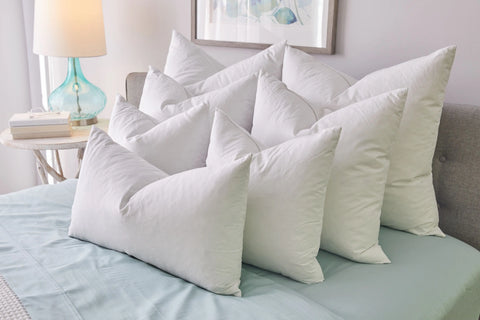 A neatly made bed features eight large, white Down Decor Pillow Inserts (Set of 2) stacked in two rows. The bed has light blue sheets, with a bedside table holding a lamp and books beside it.