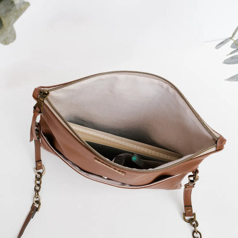 The DREAMER foldover bag in HAZELNUT by Carry Courage is open, showing a spiral notebook and essentials inside. This brown purse features a strap with brass hardware, set on a white background with green leaves in view.
