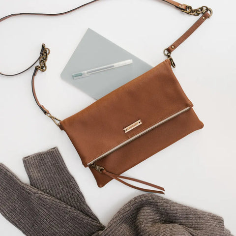 The DREAMER foldover bag in Hazelnut by Carry Courage, made of brown vegan leather with a gold zipper and detachable strap with gold hardware, is set on a white surface beside a gray notebook, white pen, and ribbed gray sweater.