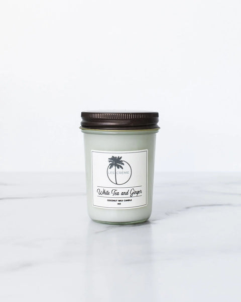 The Les Creme White Tea + Ginger Scent Coconut Wax Candle, featuring an elegant glass jar with a dark lid, sits on a white marble surface against a plain background—an inviting, toxin-free candle with an oriental fragrance.