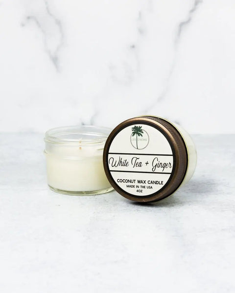 A small white, toxin-free Les Creme White Tea + Ginger Scent Coconut Wax Candle sits in a glass jar on a white surface. The 4oz lid, placed beside it, bears a palm tree logo and Made in the USA label.