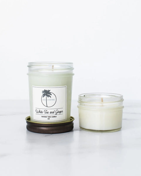 Two glass jar candles on a white surface: the taller, with light green wax, is Les Creme’s White Tea + Ginger Scent Coconut Wax Candle; the shorter, off-white candle has no visible label.