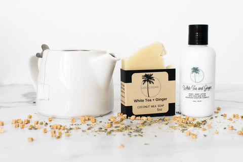 A white teapot, a bar of white tea and ginger coconut milk soap, a bottle of white tea and ginger lotion, and a Les Creme White Tea + Ginger Scent Coconut Wax Candle are arranged on a white surface with tea leaves and ginger pieces.