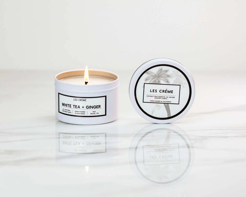 A lit Les Creme White Tea + Ginger Scent Coconut Wax Candle sits in a tin on a marble surface beside its closed lid featuring the Les Creme logo and a palm tree graphic.